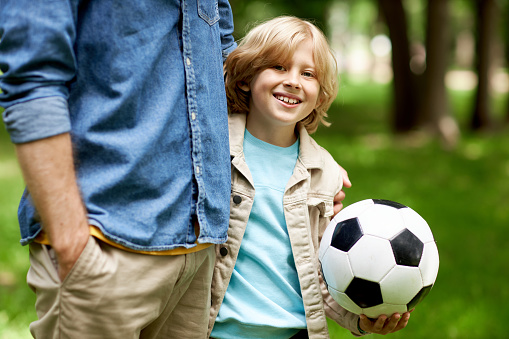 Close up of blonde boy holding football ball with father outdoors and looking at camera