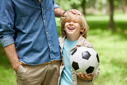 Blonde little boy holding football ball with father ruffling his hair outdoors