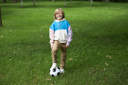 Full length portrait of smiling tween boy with football ball outdoors standing on green grass, copy space