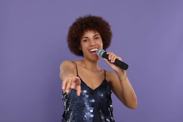 istock Curly young woman with microphone singing on purple background 1646941580