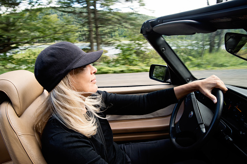 Beautiful mature woman driving convertible car in country. She is in her late fifties, dressed in black, with mid-long blonde hair, and eyeglasses. Car is a black vintage convertible. Horizontal waist up outdoors shot with copy space. This was taken in the Laurentides forest, in Quebec, Canada.