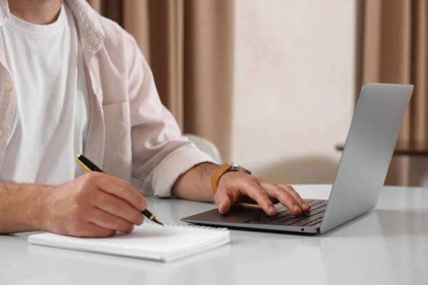istock Man using laptop and writing something in notebook at table indoors, closeup 1646978104
