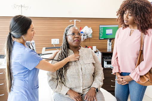 The mid adult daughter waits as the mid adult female nurse uses a stethoscope to check her mature adult mother's heart and lungs.