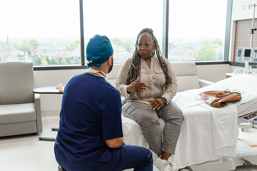 The mature adult female patient takes a chance to ask the unrecognizable male anesthesiologist her questions when he comes to see her in her hospital room before the procedure.