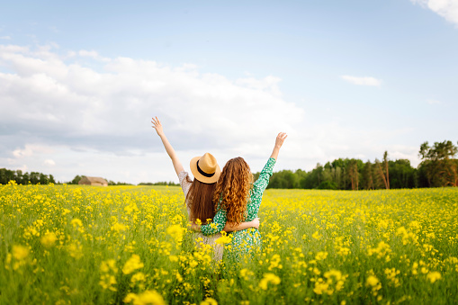 Girlfriends dressed in stylish dresses are walking together in a rapeseed field. Two women enjoy the weather, pose, have fun in a field among blooming rapeseed.