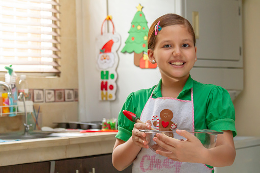 Close portrait of happiness latin young girl cooking Christmas Cupcakes for Christmas night / breakfast inside latin home in Latin America.

She's wearing a Christmas shirt. She's mixing flour and cream, on back, a nice Christmas draws did by herself.

Christmas and New Year celebration traditional treats. People cooking food.

Christmas Home Latin Experience in December Holidays.