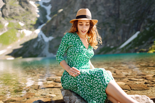 Young woman in a dress and hat enjoys the scenery on a high mountain lake. Romantic tourist posing against the background of mountain landscapes.