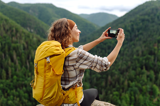 Young traveler with a yellow backpack is resting and taking pictures on the phone on a mountain cliff, admiring the landscape of the high Tatras, mountain range. Beautiful woman on top of the mountain