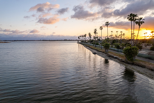 Magical view of many palm trees, the calm, shiny water of Mission Bay on Fiesta Island in San Diego, California, and the beautifully colorful sky above the bay as the sun is setting