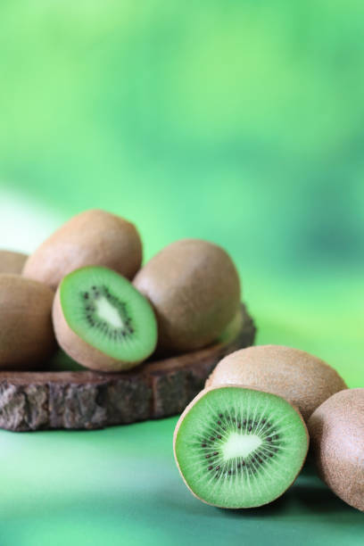 Stock photo showing a close-up view of healthy eating image of wooden fruit bowl piled with Chinese gooseberry (kiwi), one cut in half displaying bright green flesh with ring of black seeds. imagen de pila de kiwis (actinidia chinensis) en frutero de madera, grosella espinosa china cortada por la mitad mostrando piel marrón difusa y carne verde brillante con anillo de semillas negras, fondo verde moteado, enfoque en primer plano, espacio de c