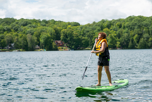 Elementary age boy wearing a life jacket and bathing suit is standing up on a SUP Stand Up Paddleboard while at a kids summer camp. He is floating in the lake with trees behind him.