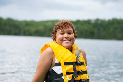 Elementary age boy with curly red hair is smiling at the camera while sitting by the lake at a cottage in a yellow life jacket for safety to help him swim.