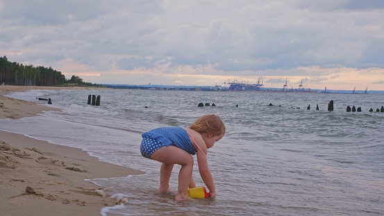 Cute Little Caucasian Toddler Girl Wearing Swimsuit Playing on Beach Trying to Fill Toy Watering Can with Water from Sea Waves Breaking on Seashore