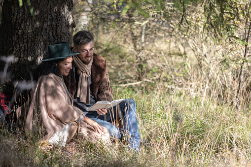 A Latin American woman and a Caucasian man storyteller, happy, in love and smiling are sitting on a date leaning against the trunk of a big tree in the grass in autumn and having fun reading a book. They are leaning on each other. Portrait.