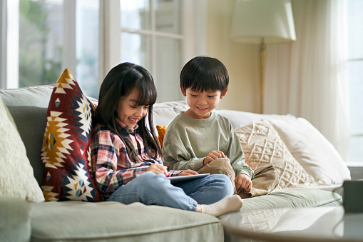 little asian children brother and sister sitting on family couch at home using digital tablet computer together