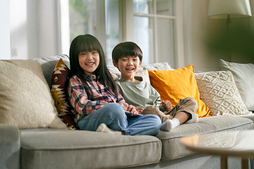 portrait of two asian children brother and sister sitting on family couch at home looking at camera smiling