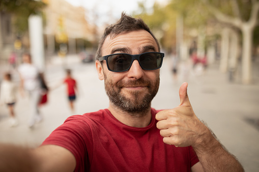 A young, bearded man, wearing sunglasses, smiles at the camera, while taking a selfie with his cell phone during a walk in the city.