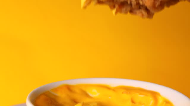 fried chicken pieces dipped in yellow cheese sauce, rotation. girl dipped crispy chicken wing in cheese sauce on a yellow background.
