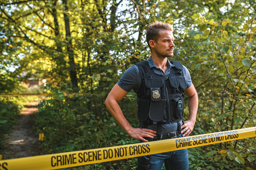Mid adult policeman in civil clothes and protective vest, standing with arms akimbo at a scene of a crime in a nature reserve. Looking away, 3/4 length image.