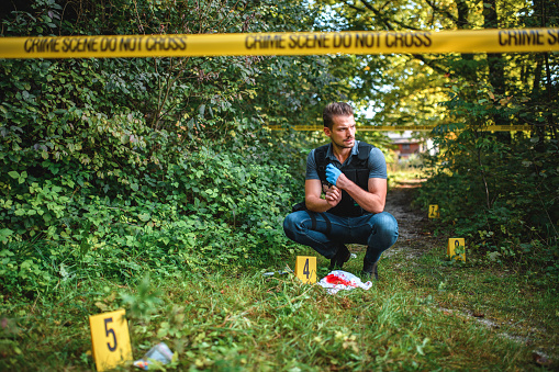 Stern mid adult forensic detective looking into the evidence at a crime scene in the forest. Crouching down with elbows leaning on his thighs, looking in the distance, taking off blue latex gloves. Full length image.