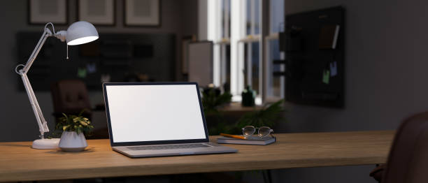 istock Dark workspace in the office at night, a laptop white screen mockup on a wooden table. 1647553132