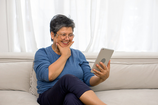 Elderly lady engrossed in digital tablet usage while relaxing on her living room settee.
