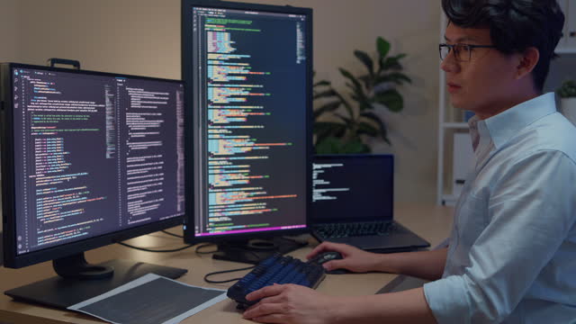 Young Asian man software developers using computer to write code sitting at desk with multiple screens work in office at night. Programmer development.