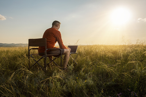 man traveler blogger work remote on netbook computer while enjoying beautiful nature landscape view outdoors at sunset.