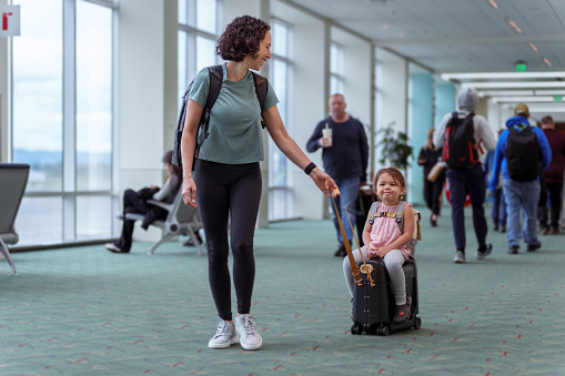 Preschool aged girl of Eurasian, Hawaiian and Pacific Islander ethnicities smiles as she sits on a suitcase, her mother pulling her through an airport.