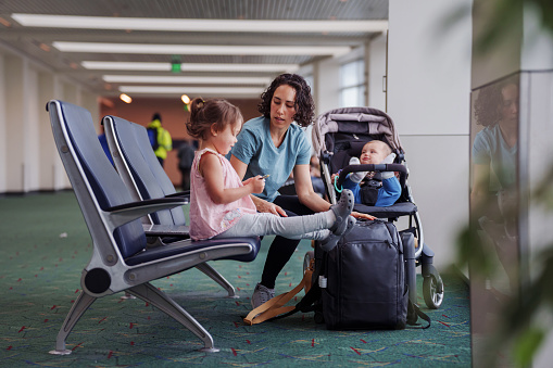 Eurasian woman with a 9 month old son talks with her three year old daughter while waiting in an airport terminal to board their flight.