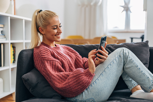 Blonde woman is sitting on the sofa and using her smart phone