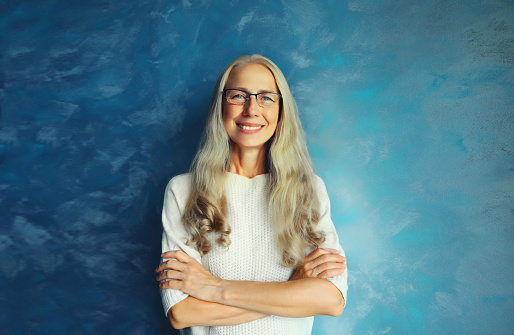 Portrait of happy smiling confident middle-aged woman with crossed arms in eyeglasses looks at the camera. Gray-haired female in casual on blue background