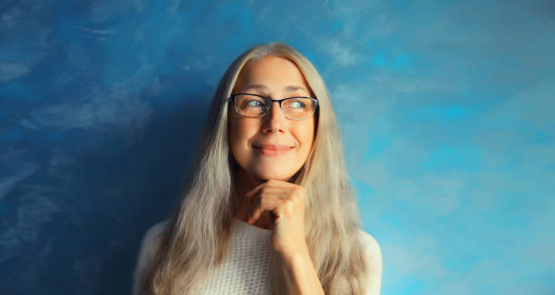 portrait d’une femme d’âge moyen souriante et souriante et souriante portant des lunettes qui détourne le regard. pensée féminine aux cheveux gris sur fond bleu - women common 40s candid photos et images de collection