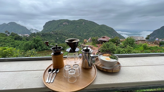 Breakfast set of drip coffee and porridge with beautiful scenic view of valley and misty mountain background at Doi Pha Hee, Chiang Rai, Thailand.