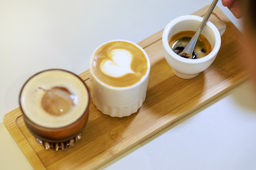 At a coffee testing event, a skillful barista places a wooden tray on the counter, artfully serving three different types of coffee. The tray holds three cups, each containing a unique coffee variant - espresso, latte, and ice black coffee.