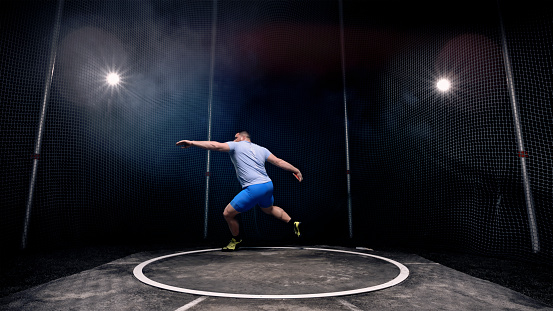 Male athlete turning with discus while practising on throwing circle at night.