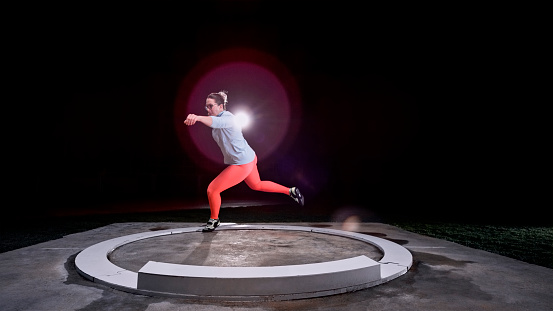 Female shot put athlete standing on one leg while throwing the metal ball at night.