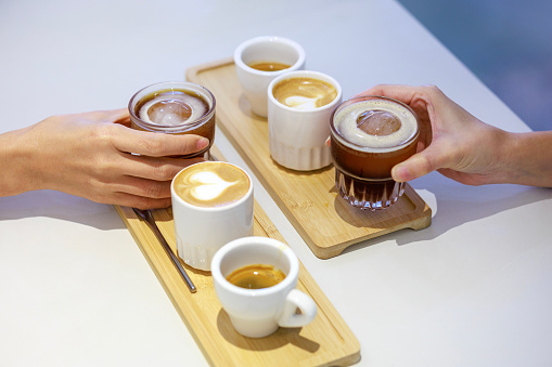 At a coffee testing event, a skillful barista places a wooden tray on the counter, artfully serving three different types of coffee. The tray holds three cups, each containing a unique coffee variant - espresso, latte, and ice black coffee.