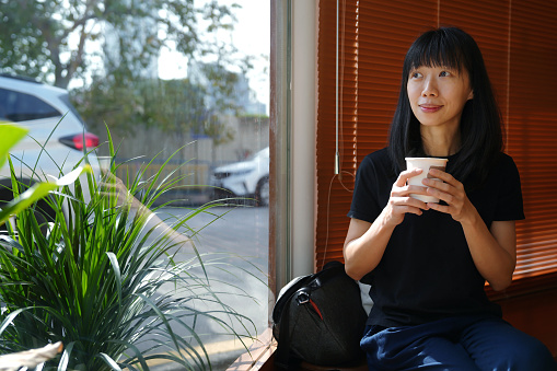 An Asian tourist woman holding a cup of coffee by the window in a cafe, she is enjoying the view outdoors