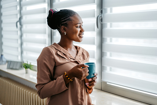 Beautiful young black woman standing by the window at home and drinking coffee.