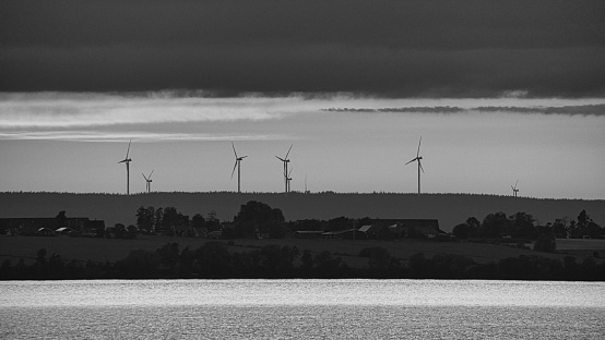 Wind turbine, renewable energy, on a lake at sunset taken in black and white. Clean electricity for climate change. Cloudy sky with orange sun in Scandinavia.