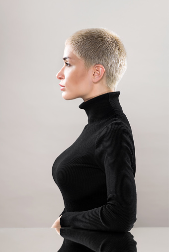 Side view of concentrated young female with short blond hair in black turtleneck sitting at mirrored table and looking away with confidence against beige background