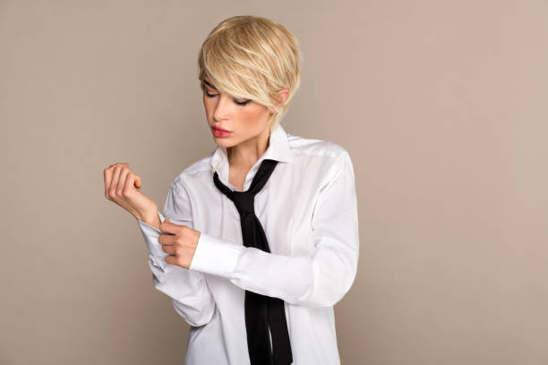 Young woman standing and buttoning shirt sleeve near beige colored wall Focused young female blondie in white shirt and tie looking down while standing and putting button on full sleeve with hand during dressing against beige background woman necktie stock pictures, royalty-free photos & images