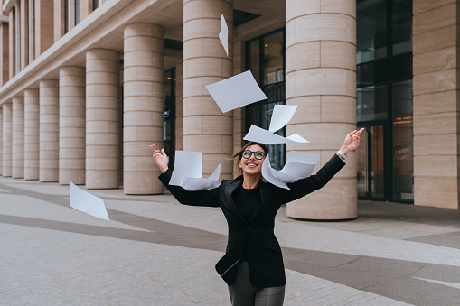 Happy American female lawyer in business suit throwing up in the air sheets of paper, smiles wide after successful end of issue. Excited businesswoman celebrating great deal. Success, happiness.