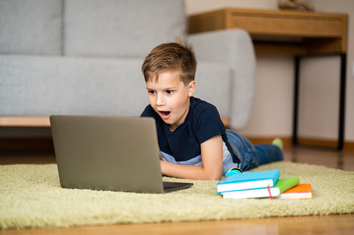 Shocked boy lying on the carpet and looking at laptop at home