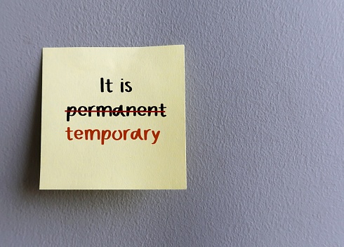 Sticky note on copy space wall with handwritten text IT IS PERMANENT crossed off to IT IS TEMPORARY, concept of optimist vs pessimist different thinking when bad things happen