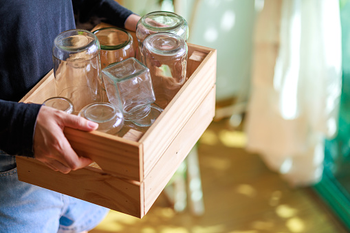 Cropped image of an Asian woman's hands placing empty glass jars into a wooden box, ready for recycling, showcasing a sustainable lifestyle.
