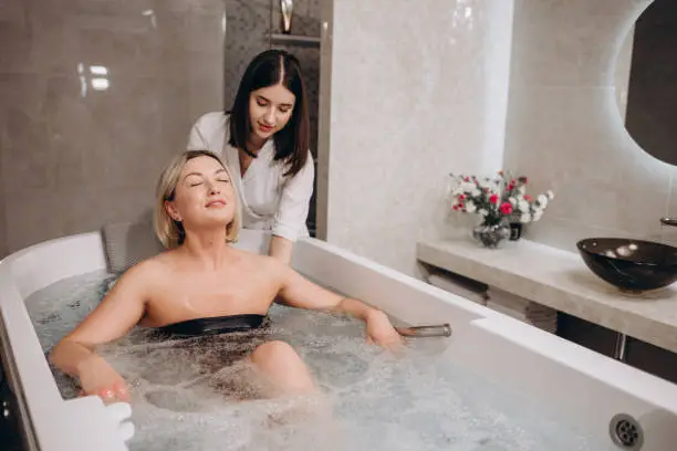 Portrait of cheerful young woman in swimsuit relaxed sitting in bath with hydromassage at luxury spa salon, looking at camera. Happy female lying in bathtub relaxing hydrotherapy in beauty center.