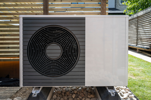 Front view of the air source heat pump outside in the garden, near wooden fence on a sunny day.