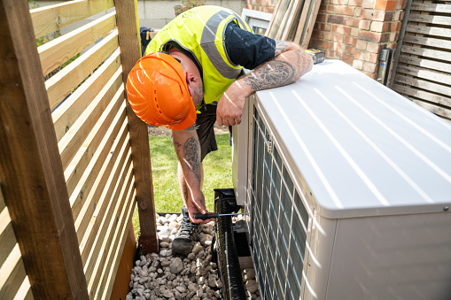 A male electrician installing a heat pump in a garden on a sunny day.